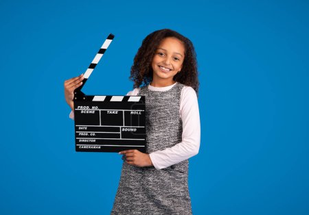 Téléchargez les photos : Heureuse adolescente afro-américaine applaudissant clapperboard pour commencer à tourner des vidéos isolées sur fond de studio bleu. Blog, enfance, production vidéo, film, cinéma avec enfant - en image libre de droit