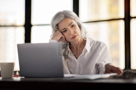 Photo for Stressed Middle Aged Businesswoman Using Laptop And Looking Through Papers Working In Modern Office. Female Entrepreneur Doing Paperwork Near Computer At Workplace. Business Documentation Problems - Royalty Free Image