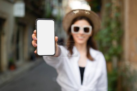 Foto de Alegre joven millennial mujer caucásica turista en gafas de sol, sombrero muestran teléfono con pantalla en blanco en la ciudad. Aplicación para la aventura, sitio web de recomendación, vacaciones y viajes blog al aire libre, mapa en línea - Imagen libre de derechos
