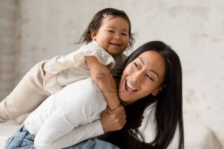 Photo for Cheerful Asian Mother Riding Adorable Little Baby Daughter On Back Laughing And Having Fun Playing In Bedroom At Home. Mommy Cuddling With Child Girl. Maternity And Babysitting Concept - Royalty Free Image