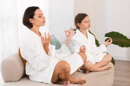 Photo for Zen. Peaceful Young Mom And Little Daughter In Bathrobes Meditating Together At Home, Calm Beautiful Mother And Cute Female Child Sitting On Sofa With Eyes Closed, Practicing Yoga In Lotus Position - Royalty Free Image