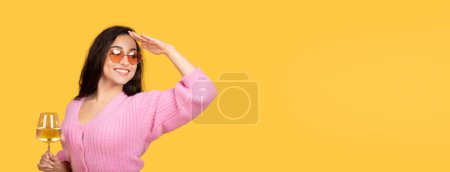 Téléchargez les photos : Glad millénaire arabe femelle dans les lunettes de soleil tenir un verre de vin léger, regarder l'espace de copie isolé sur fond jaune, studio, panorama. Fête, félicitations, alcool et détente - en image libre de droit