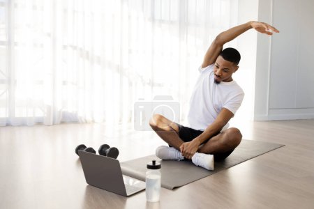 Photo for Sporty young black man in sportswear doing yoga at home, stretching body on fitness mat and watching online fitness video on laptop, training in living room, have morning workout, copy space - Royalty Free Image