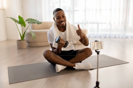 Photo for Happy smiling handsome athletic young black man pumping isron, recommending nice fitness mobile application, sitting on yoga mat with dumbbell at home, looking at cell phone screen, showing thumb up - Royalty Free Image