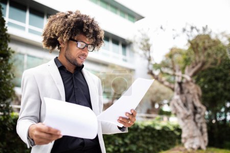 Photo for Confident african american male entrepreneur in formal outfit holding documents or CV, getting ready for job interview, standing outdoors, copy space - Royalty Free Image