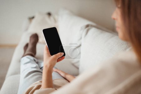 Photo for Serious millennial european female typing on phone with empty screen, sits on sofa, resting, enjoy spare time in minimalist living room interior, cropped. Social media chat, app at home - Royalty Free Image