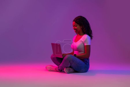 Téléchargez les photos : Travail à distance. Femme noire souriante utilisant un ordinateur portable dans la lumière au néon, heureuse jeune afro-américaine pigiste dame assise sur le sol avec ordinateur, posant sur dégradé fond violet, Copier l'espace - en image libre de droit