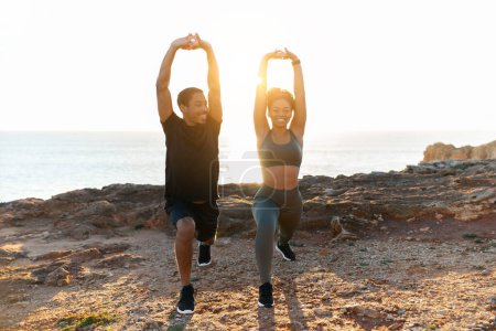 Photo for Glad young african american female and man in sportswear doing stretching, practice yoga, enjoy weight loss on ocean beach at sunrise. Body care and health care together, morning workout outdoor - Royalty Free Image