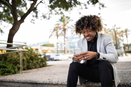 Téléchargez les photos : Entrepreneur noir d'âge moyen qui réussit à utiliser un téléphone portable, envoyer des SMS ou naviguer sur Internet, assis sur des escaliers à l'extérieur, faisant une pause dans le parc, espace libre - en image libre de droit