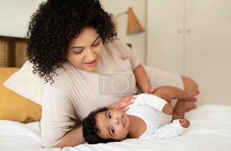 Téléchargez les photos : Soins aux bébés, parentalité et maternité. Souriant jeune afro-américaine femelle et petit enfant allongés sur le lit, se détendre ensemble, jouer à l'intérieur de la chambre. Amour, relation maman et enfant à la maison - en image libre de droit