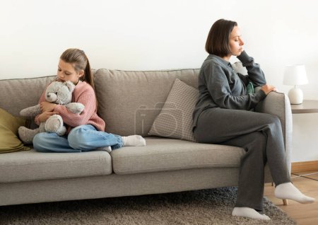 Sad offended european millennial mom ignores teen girl, sit on sofa in living room interior after quarrel. Stress, scandal, teenage issues and relationships problems at home