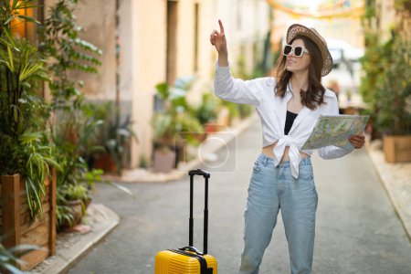 Photo for Serious young millennial caucasian lady tourist in hat with map and yellow suitcase points finger at empty space in city. Weekend, adventure vacation and travel alone outdoor - Royalty Free Image