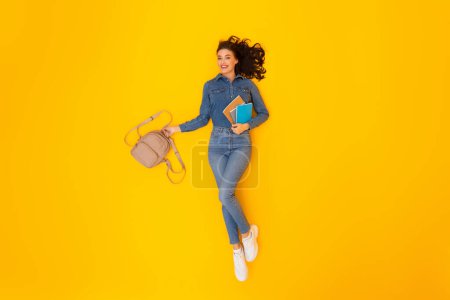 Photo for Cheerful Student Lady Holding Backpack And Copybooks Posing Lying Over Yellow Studio Background, Smiling To Camera. Modern Education And Knowledge Concept. Above View Shot - Royalty Free Image