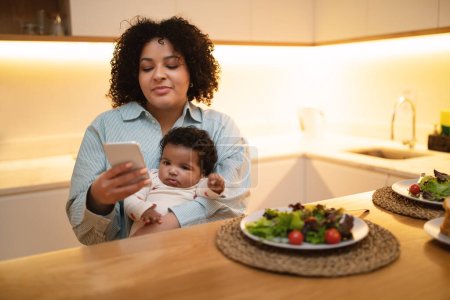 Foto de Sonriente joven latina rizada madre sentada en la mesa de la cocina, sosteniendo al bebé toddle en su regazo mientras almuerza, usando un teléfono inteligente, mujer de negocios trabajando remotamente mientras cuida a su hijo en casa - Imagen libre de derechos