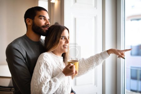 Photo for Cheerful millennial arabic husband embracing caucasian wife with cup of tea, point finger to window enjoy romantic, free time in living room interior. Coffee break, love, relationship, family home - Royalty Free Image