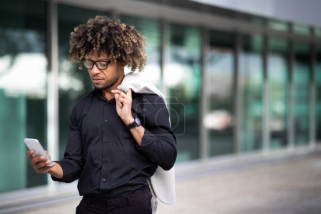 Photo for Handsome black middle aged businessman using cellphone, texting with client or surfing internet, holding jacket on shoulder, walking outdoor near business center, free space - Royalty Free Image