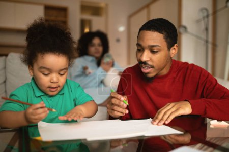 Photo for Loving handsome young african american man in casual father spending time with his kid at home, drawing with his little adorable son, woman mother feeding infant baby on background - Royalty Free Image