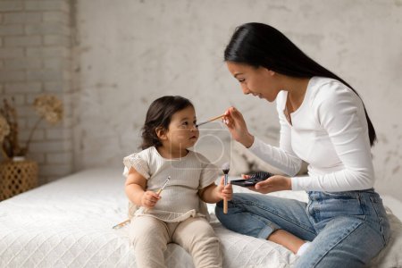 Photo for Happy Asian Mother And Baby Having Fun With Cosmetics Touching Daughters Nose With Brush Making Makeup Sitting On Bed In Modern Bedroom At Home. Mommy And Child Playing With Beauty Products - Royalty Free Image
