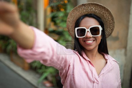 Foto de Videollamada, rodaje blog de viajes. Feliz joven millennial turista negro en sombrero y gafas de sol tomando selfie en el teléfono, disfrutar de la aventura en la ciudad, de cerca. Aplicación para vacaciones solo al aire libre y viaje - Imagen libre de derechos