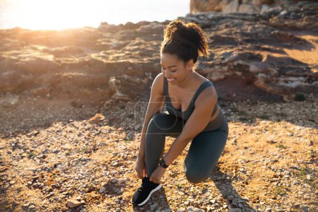 Photo for Positive millennial african american female in sportswear tying shoelaces on sneakers, on ocean beach at sunrise. Body care and health care, morning workout, weight loss outdoor - Royalty Free Image