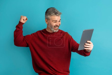 Téléchargez les photos : Pari en ligne, jeu. Positif heureux beau cheveux gris homme d'âge moyen en rouge serrant le poing, levant la main vers le haut, mâle célébrant le succès, tenant tablette numérique, fond bleu studio - en image libre de droit