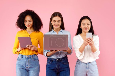 Téléchargez les photos : Trois femmes multiraciales utilisant des gadgets et regardant des écrans, naviguant sur Internet sur fond de studio rose. Réseau de groupes de femmes dans les médias sociaux en ligne - en image libre de droit
