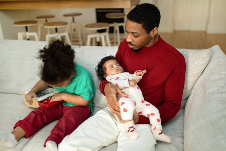 Photo for Young handsome black man in casual father spending time with his little kids at home, dad sitting on couch, carrying sleeping little baby girl, watching his son toddler playing with toy - Royalty Free Image