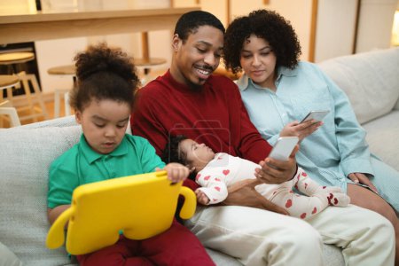 Téléchargez les photos : Happy famille multiculturelle aimante père noir, mère hispanique, deux petits enfants assis sur le canapé à la maison, en utilisant des gadgets, homme et femme tenant smartphones, tout-petit regardant bande dessinée sur tablette numérique - en image libre de droit