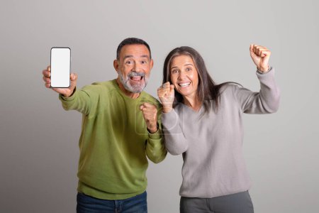 Téléchargez les photos : Heureux vieil homme et femme européens excités montrent smartphone avec écran blanc, se réjouir du succès, célébrer la victoire sur fond de studio gris. Victoire en ligne, émotions des gens et expression faciale - en image libre de droit