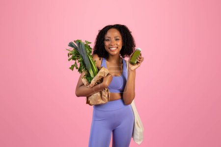 Smiling millennial black female in sportswear show paper bag with green vegetables and smoothie bottle isolated on pink studio background. Weight loss, body and health care, vegetarian food