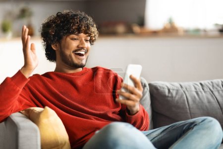 Photo for Good News. Happy Young Indian Man Looking At Smartphone Screen While Sitting On Couch At Home, Handsome Eastern Male Raising Hand And Exclaiming With Excitement, Celebrating Online Win - Royalty Free Image