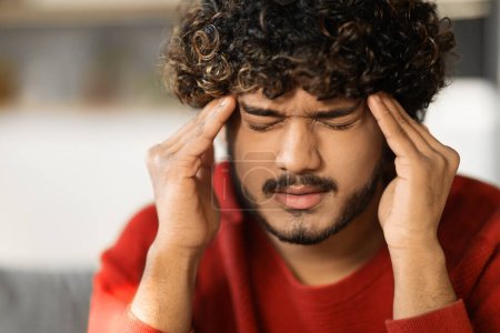 Photo for Closeup Shot Of Stressed Young Indian Guy Suffering From Headache At Home, Millennial Eastern Man Sitting On Couch, Frowning And Touching Temples, Having Acute Migraine, Feeling Unwell - Royalty Free Image