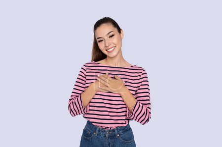 Photo for Cheery happy thankful pretty hispanic young woman wearing casual outfit holding hands on chest and smiling at camera over grey studio background, expressing love, affection, copy space - Royalty Free Image