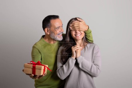 Foto de Feliz viejo hombre europeo cierra los ojos a la mujer, da caja de regalo sobre fondo gris estudio. Celebración de vacaciones, felicitaciones, aniversario de cumpleaños, emociones de la gente y sorpresa, aniversario - Imagen libre de derechos