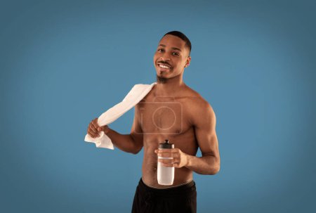 Photo for Sports lifestyle. Happy handsome shirtless young black guy sportsman with towel on his shoulder and bottle of water in his hand isolated on blue studio background, smiling at camera, copy space - Royalty Free Image
