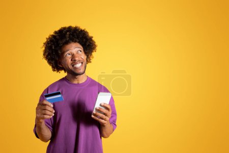 Téléchargez les photos : Joyeux cher adulte africain américain bouclé homme en t-shirt violet avec smartphone et carte de crédit regarde l'espace vide, pense à l'ordre sur fond de studio jaune. Achats en ligne, banque - en image libre de droit