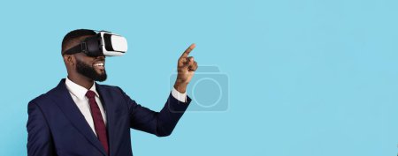 Téléchargez les photos : Technologies modernes pour les entreprises. Portrait d'un homme d'affaires noir souriant portant un casque VR, un homme d'affaires afro-américain heureux jouant à un jeu vidéo et pointant du doigt sur un terrain noir bleu - en image libre de droit