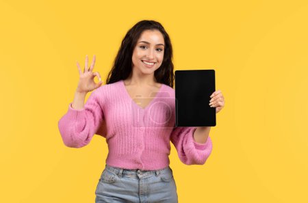 Photo for Positive millennial arab lady in casual show tablet with empty screen, ok sign with hand isolated on yellow background, studio. Recommendation, gadget presentation, advice website and app - Royalty Free Image