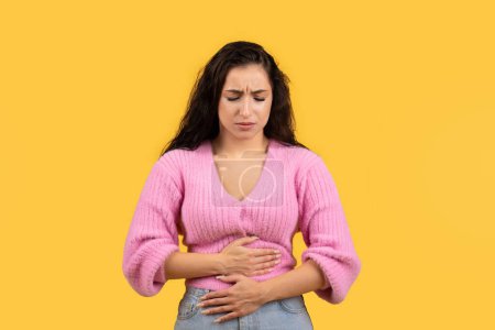 Photo for Sad millennial arab lady presses her hands to belly, suffering from stomach pain, health problems isolated on yellow background, studio. Ulcer, disease and poisoning, diarrhea, menstruation - Royalty Free Image