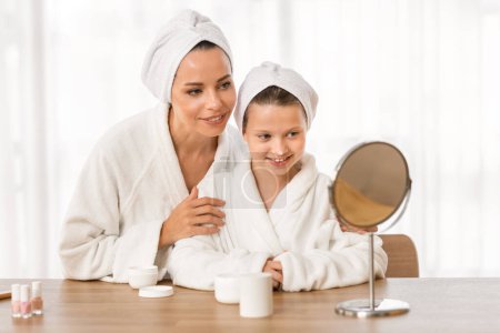 Photo for Beautiful mother and daughter in bathrobes and towels applying face cream together, happy mom and cute preteen female child sitting at dressing table and looking in mirror, enjoying beauty routine - Royalty Free Image
