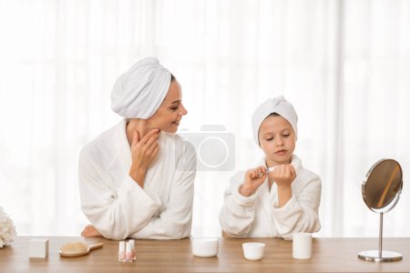 Téléchargez les photos : Petite fille mignonne remplissant des ongles tout en ayant une journée spa avec maman, souriant Preteen enfant féminin et sa mère portant des peignoirs de bain jouant salon de beauté à la maison, profiter de passer du temps ensemble - en image libre de droit