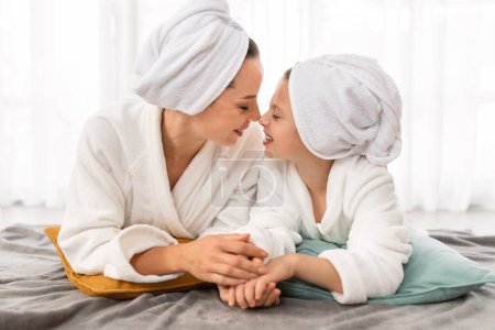 Photo for Mothers Day Concept. Mom And Little Daughter Wearing Bathrobes Relaxing On Bed Together, Loving Mommy And Cute Female Child Touching Noses And Smiling, Bonding At Home, Closeup Shot - Royalty Free Image