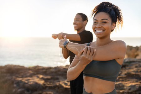 Photo for Smiling millennial african american lady and male in sportswear doing arm stretching, enjoy morning workout on ocean beach at sunrise. Body care and health care together, weight loss outdoor - Royalty Free Image