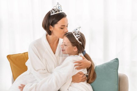 Photo for Mothers Love. Loving Mom Kissing Her Cute Little Daughter While They Relaxing In Bathrobes At Home, Happy Mommy And Female Child Wearing Toy Crowns, Enjoying Spending Time Together, Copy Space - Royalty Free Image