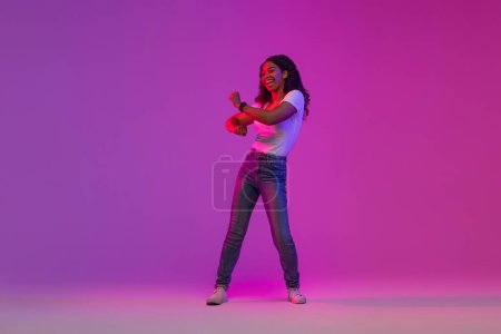 Téléchargez les photos : Jeune femme noire joyeuse dansant dans la lumière au néon sur fond violet, pleine longueur prise de vue de heureuse femme afro-américaine millénaire positive s'amusant dans le studio, profiter de la fête, copier l'espace - en image libre de droit