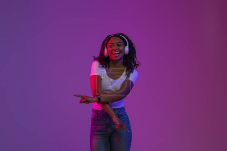 Foto de Joyful Young Black Lady en auriculares inalámbricos escuchando música y bailando, Happy Carefree African American Woman disfrutando de su lista de reproducción favorita, de pie en luz de neón sobre fondo púrpura - Imagen libre de derechos