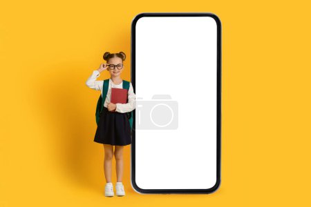 Photo for Excited Little Schoolgirl Standing Near Big Blank Smartphone With White Screen, Cute Smiling Preteen Female Child Advertising Educational App Or Website, Posing On Yellow Background, Mockup - Royalty Free Image