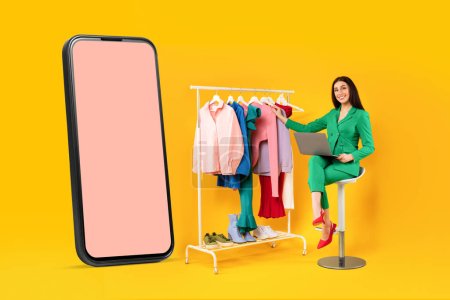 Téléchargez les photos : Heureuse femme accro au shopping assise près d'un grand téléphone portable et d'un rail de vêtements, choisissant et achetant des vêtements en ligne, une maquette, un fond de studio jaune - en image libre de droit