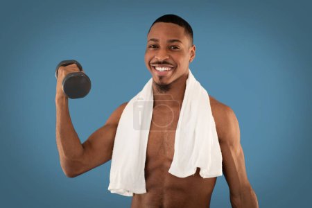 Photo for Cheerful handsome athletic young black man with towel on his shoulders exercising with barbell on blue studio background, african guy working on arms strength, looking at camera and smiling - Royalty Free Image