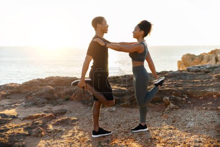Téléchargez les photos : Couple afro-américain millénaire positif en vêtements de sport faisant étirement pour les jambes, pratiquer le yoga sur la plage de l'océan au lever du soleil, profil. Entraînement matinal, soin du corps ensemble, remise en forme extérieure - en image libre de droit